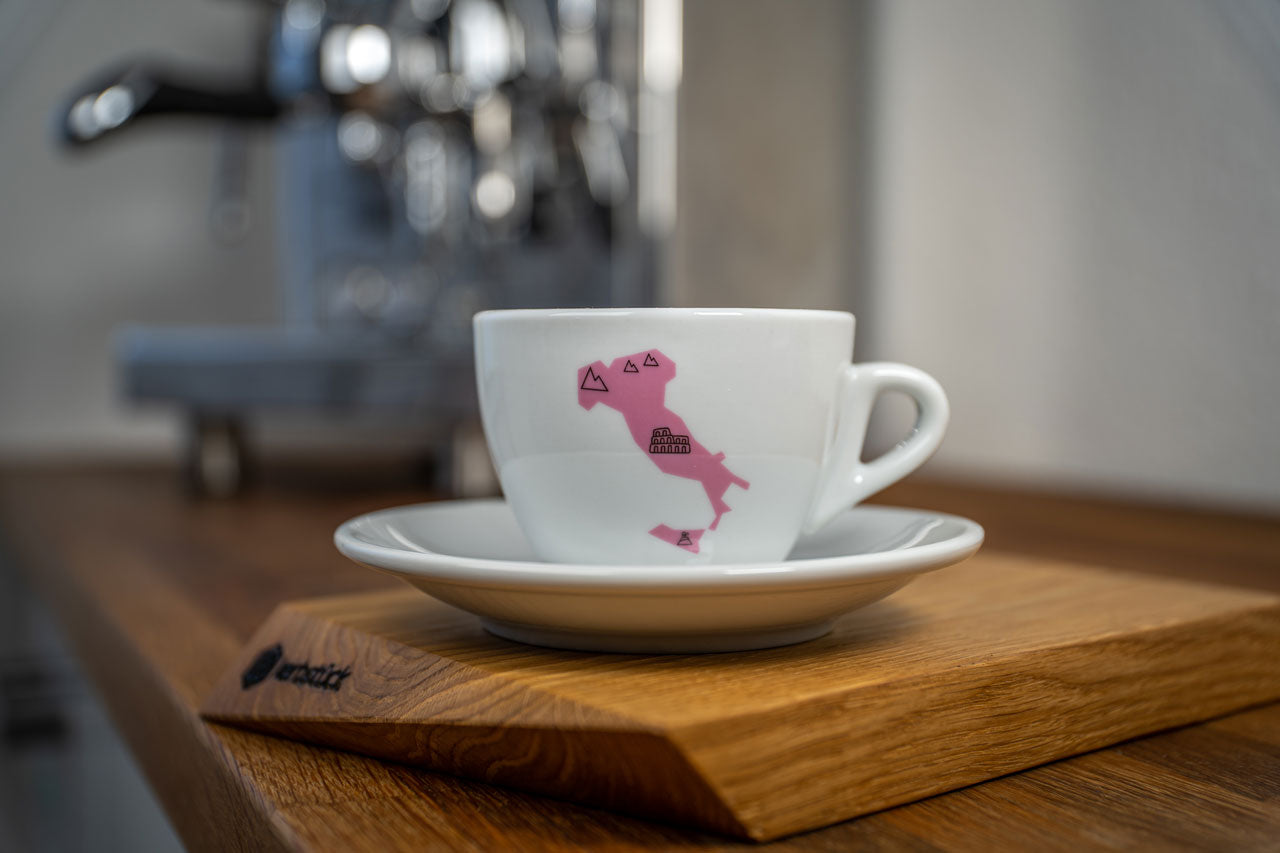 Eine weiße Cappuccino-Tasse bedruckt mit der rosanen Silhouette von Italien. In schwarz darauf das Kolosseum und Symbole für Berge und Wind.