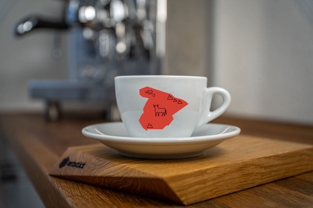 Eine weiße Cappuccino-Tasse bedruckt mit der roten Silhouette von Spanien. In schwarz darauf ein Stier und Symbole für Berge und Wind.