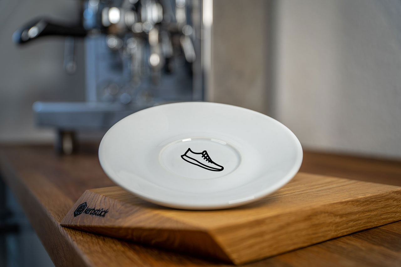 Eine weiße Cappuccino-Untertasse bedruckt mit einem Icon eines Laufschuhes. Im Hintergrund eine glänzende Siebträger-Kaffeemaschine.