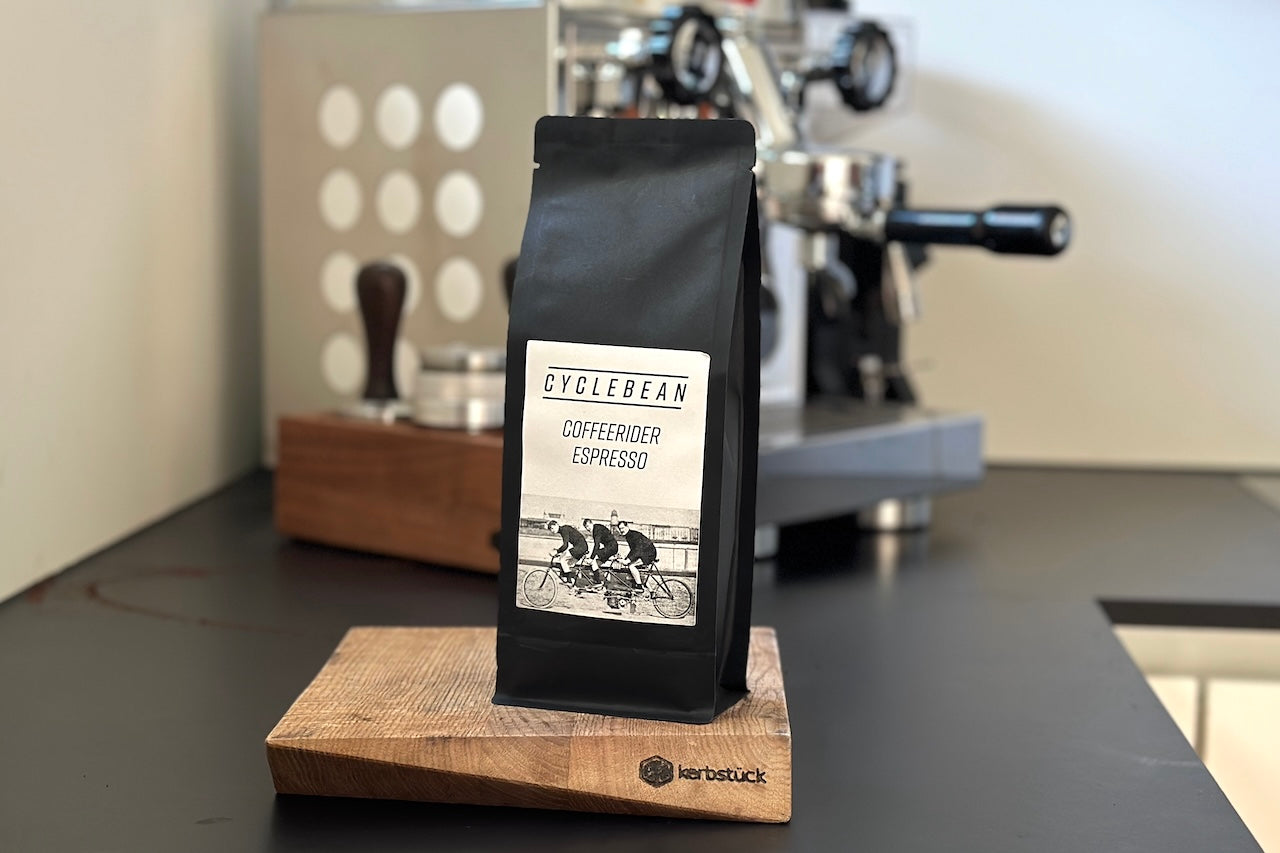 Eine Espresso-Packung steht auf einer Arbeitsfläche vor einer Kaffeemaschine. Auf den Label sind drei Radfahrer zu sehen.