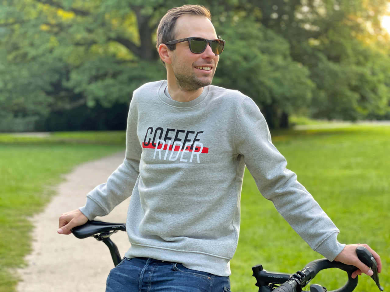 Eine Person steht in einem Park und trägt einen grauen Pullover mit der Aufschrift Coffee Rider