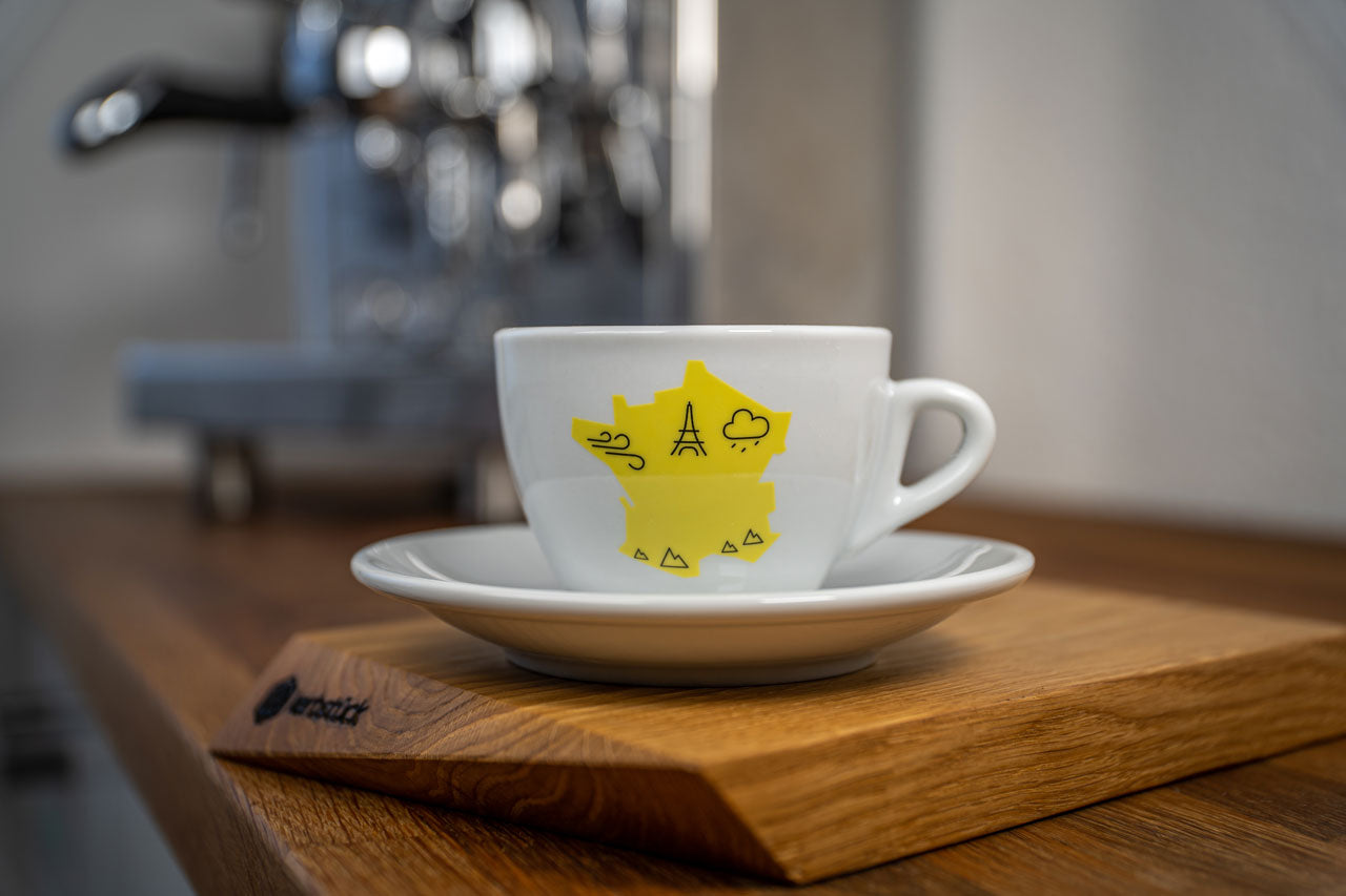 Eine weiße Cappuccino-Tasse bedruckt mit der gelben Silhouette von Frankreich. In schwarz darauf der Eifelturm und Symbole für Berge, Wind und Regen.