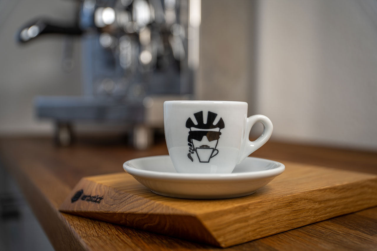 Eine weiße Espresso-Tasse bedruckt mit einem stilisierten Kopf einer Radfahrerin mit Helm, Sonnenbrille und Zopf, die aus einer Kaffeetasse trinkt.