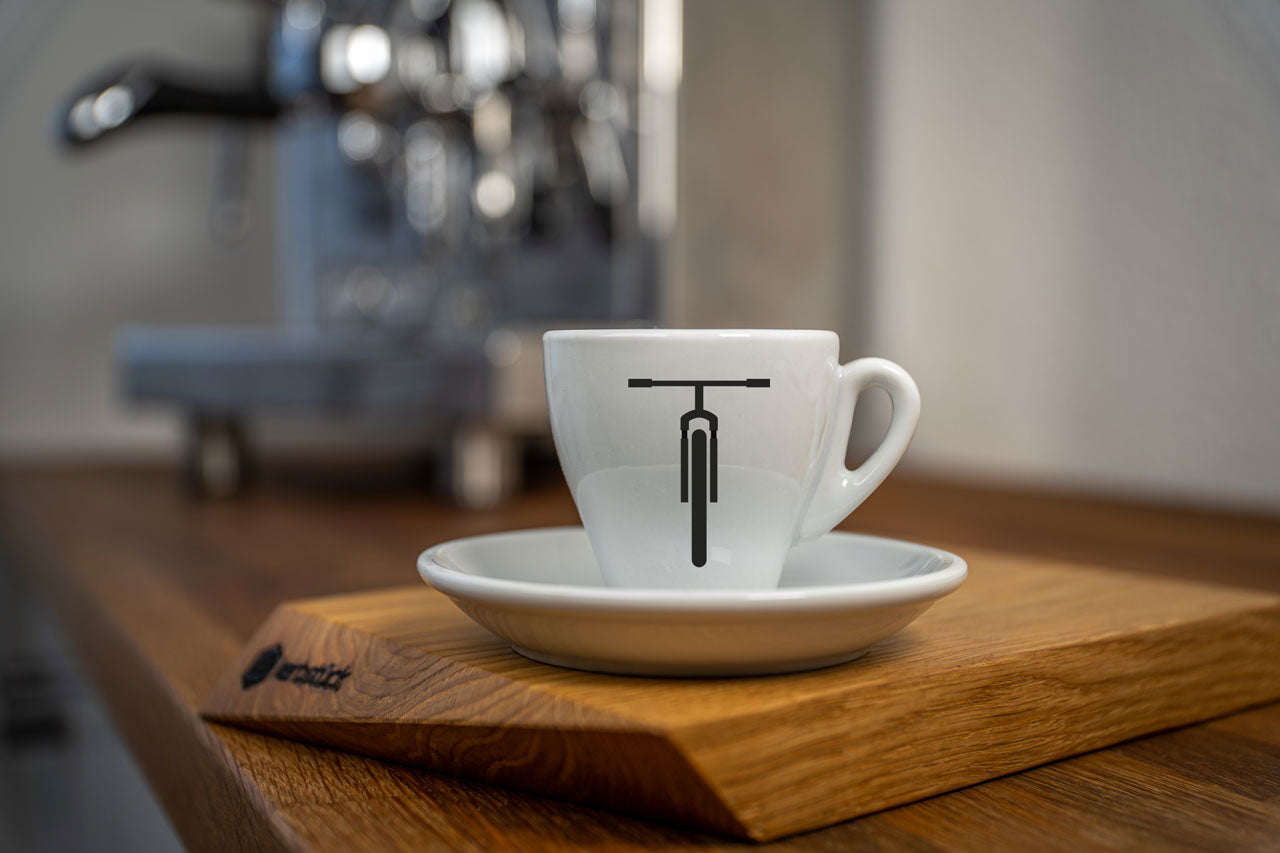 Eine weiße Espresso-Tasse bedruckt mit der schwarzen Moutainbike-Silhouette. Im Hintergrund eine glänzende Siebträger-Kaffeemaschine.